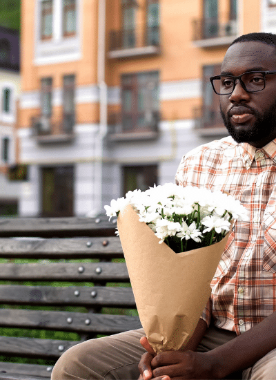 African man sitting lonely on city bench, holding flower bouquet,